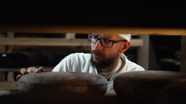 Concentrated young man checking cheese ripening beating wheel with hammer in slow motion. Front view of expert male Caucasian cheese maker in warehouse indoors. Shooting behind shelves