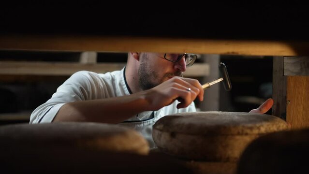 Professional cheese maker beating cheese wheel with hammer checking ripening indoors. Portrait of expert Caucasian man examining dairy product in warehouse