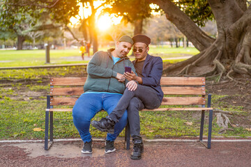 Gay Latino male couple sitting on a bench in a park at sunset, wearing fashionable hats, holding a cell phone, smiling