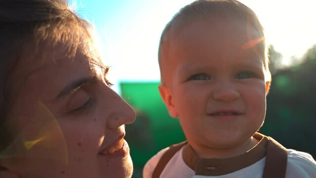 Loving mother touches tiny nose of little son holding in arms on blurred background. Brunette woman and blond toddler stand in yard closeup slow motion