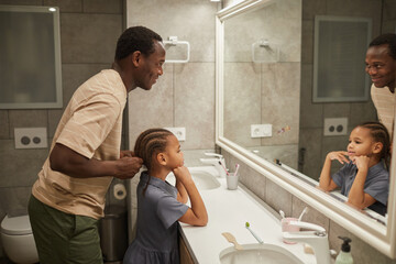 Side view portrait of caring black father braiding daughter's hair by mirror in bathroom and helping get ready in morning