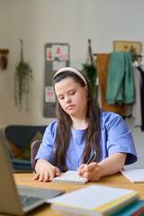 Female student with Down syndrome making notes in notepad during online lesson while sitting by desk in front of laptop at home © pressmaster