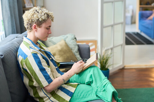 Young Female Amputee In Casualwear Making Sketches With Pencil In Notepad While Sitting On Soft Couch In Home Environment