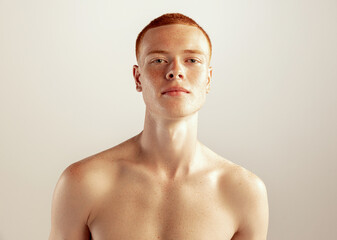 Portrait of young red-haired man posing shirtless isolated over grey studio background. Freckled...