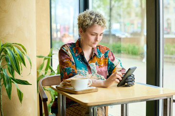 Young woman with physical impairment scrolling in smartphone while sitting by table in cafe and having coffee with sandwiches