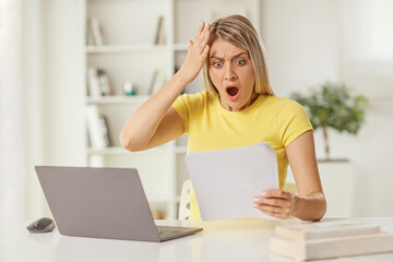 Shocked young woman looking at a document and sitting at a laptop computer