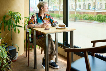 Young woman in casualwear looking through window while sitting by table in cozy cafe and having coffee with snack for breakfast