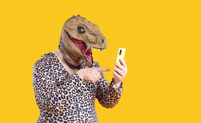 Ecentric funny fat man in dinosaur mask uses mobile applications and communicates on social networks. Chubby man with dinosaur head in leopard clothes holding mobile phone on orange background.