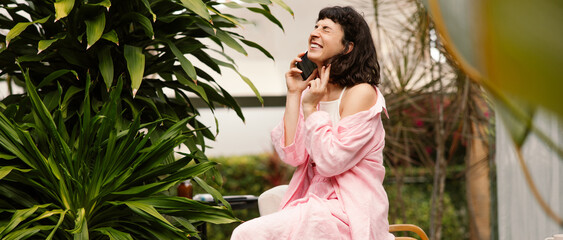 Cute young caucasian girl with closed eyes laughs talking on phone outdoors. Brunette with bob haircut wears pink suit. Emotion concept, facial expression.