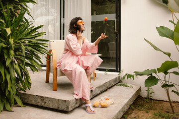 Funny young caucasian woman tosses tangerine sitting on chair outdoors during day. Brunette with short haircut wears pink suit. People sincere emotions lifestyle concept.