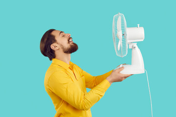Funny bearded man enjoys fresh air and escapes summer heat with electric fan isolated on light blue...