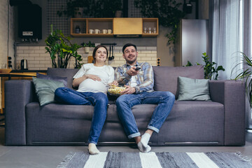 Home cinema. Young family. Pregnant woman and man sitting on sofa at home, watching TV, movie,...