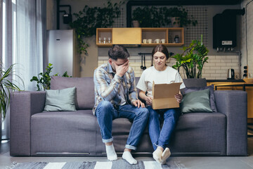 Fototapeta premium A young family, a pregnant woman and a man accidentally received someone else's parcel. They opened the box, disappointed. Sitting at home on the couch, sad. The man holds his head