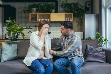 Obraz premium Young pregnant woman crying, wipes tears with a napkin. The man calms her down, supports her. Sitting at home on the couch. Emotional state of a pregnant woman