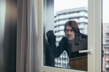 Young male robber in dark clothes looks out the window of the apartment, house. He wants to go into...