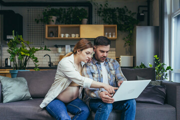 Fototapeta premium Online shopping. Young family, pregnant woman and man make an online purchase order from a laptop, holding a credit card. Sitting at home on the couch. Very happy, satisfied.