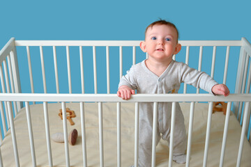Funny baby boy is standing in the crib, studio blue background. Fun child in white pajamas, copy space