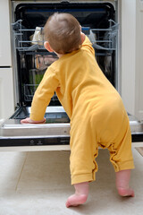 Baby boy toddler is standing by an open dishwasher with dirty dishes inside. Child safety issues in...
