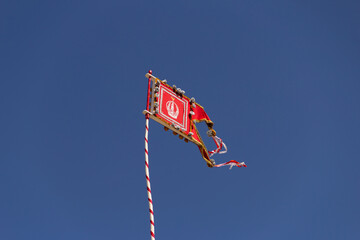 Uma flâmula do Divino Pai Eterno, presa em um pole com céu azul ao fundo.