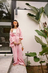 In full growth, cute caucasian brunette girl is resting outdoors standing near house. Model wears sunglasses, pink shirt and pants. People sincere emotions lifestyle concept.