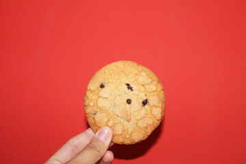 Round texture cookie in a female hand on a red background, close-up, front view. Cookie with cracks on the surface and four small pieces of chocolate. Dessert. Yummy.