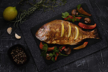 Grilled fish dorada with roasted ingredients and lemon  on dark tray. Top view. Free copy space.