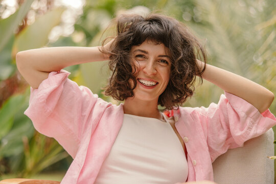 Happy young caucasian girl looking at camera holding head with her hands outdoors. Brunette woman with wavy short hair wears top and shirt. People emotions, lifestyle and fashion concept.