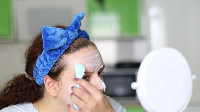Woman use spatula to apply moisture anti-wrinkle mask before facial foundation, moisturizer cream, looking in mirror. Rejuvenation. Face skincare, professional cleanser product cosmetics. Peeling skin