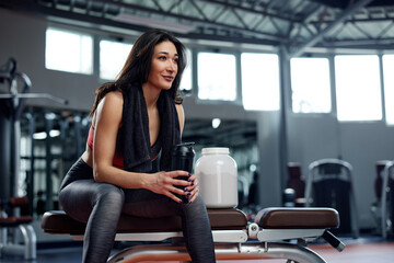 Young active woman taking a pause in the gym. She is sitting, holding a protein shake with a towel...