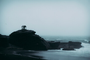 Nicho o grutita de madera con virgen dentro sobre una roca en la playa en día nublado con olas rompiendo en la costa