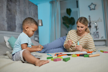 Portrait of cute black toddler playing with building blocks on bed, Caucasian mother in background, copy space