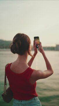 VERTICAL VIDEO: Close-up, girl stand on the embankment and takes pictures of sunrise on mobile phone. Closeup of young woman shoots the sea on smartphone in the morning time. Back view