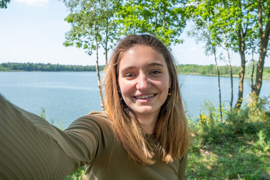 Beautiful cheerful young woman having a good time at the forest lakeside on a lovely day, taking a selfie, smiling. High quality photo