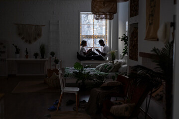 A couple of African Americans sit opposite each other on the windowsill and look at the phones.