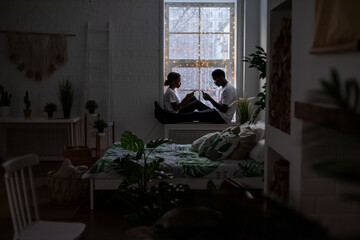 A couple of African Americans sit opposite each other on the windowsill and look at the phones.