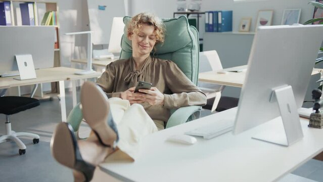 Smiling beautiful woman holding smartphone in hands, chatting. Carefree chief accountant of company relaxing in office, keeping legs on table while sitting at working place. Daytime, indoors