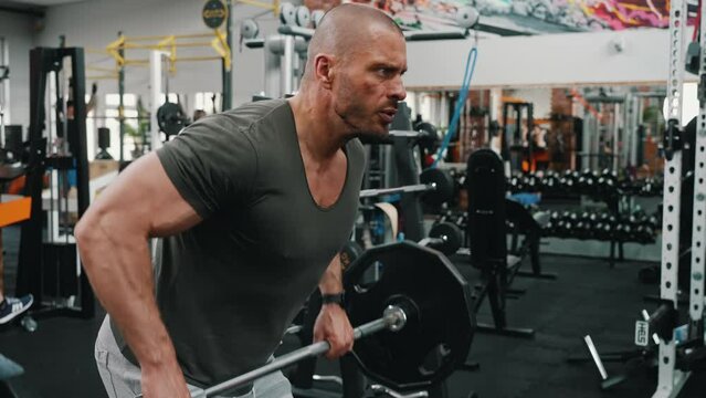 Concentrated millenial bald muscular guy lifting a heavy barbell using his triceps muscles. Indoor shot at a gym. High quality 4k footage