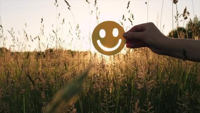 Female hand holding happy face emoticon near green grass on background amazing sunset at beautiful summer day. Positive picture or cheerful life, World mental health day and eco-friendly concept