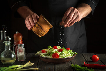 Professional chef sprinkles salted fresh vegetable salad in a plate on a wooden table. Cooking healthy food in the kitchen in a restaurant