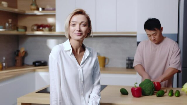 Young attractive Caucasian woman looking at man cooking. Young Asian man cutting salad, preparing breakfast in modern kitchen. Healthy relationships without gender stereotypes. True love concepts