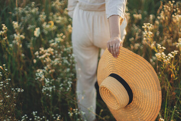 Stylish boho woman with straw hat in hand close up among wildflowers in sunset light. Atmospheric moment. Summer delight and travel. Young female in rustic linen cloth walking in summer meadow