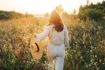 Stylish boho woman with straw hat walking among wildflowers in warm sunset light. Atmospheric...