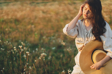 Obraz premium Stylish boho woman with straw hat posing among wildflowers in warm sunset light. Summer delight and travel. Young female in rustic linen cloth relaxing in atmospheric summer meadow. Copy space
