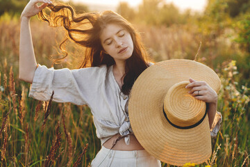 Stylish boho woman with straw hat posing among wildflowers in sunset light. Summer delight and...
