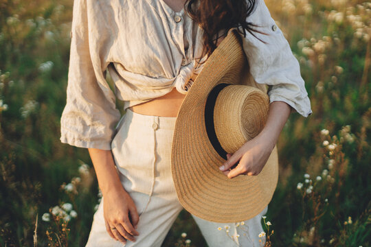 Young carefree female in rustic linen cloth close up relaxing in summer meadow. Summer delight and travel. Stylish boho woman with straw hat posing among wildflowers in sunset light