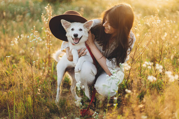 Stylish happy woman playing with cute dog with hat among wildflowers in sunset light. Summer travel...