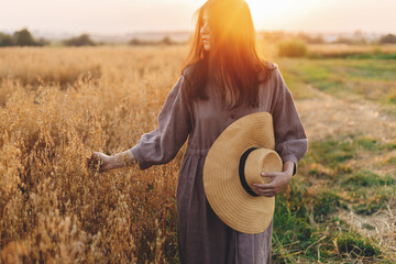 Stylish woman with straw hat walking at oat field in sunset light. Atmospheric tranquil moment....