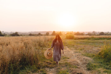 Stylish woman with straw hat dancing at oat field in sunset light. Atmospheric happy moment. Young...