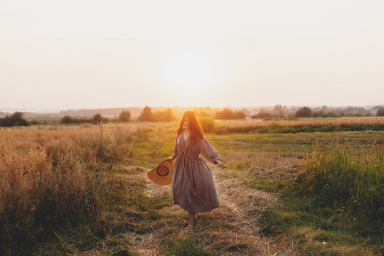 Stylish woman with straw hat dancing at oat field in sunset light. Atmospheric happy moment. Young female in rustic linen dress relaxing in evening summer countryside, rural life