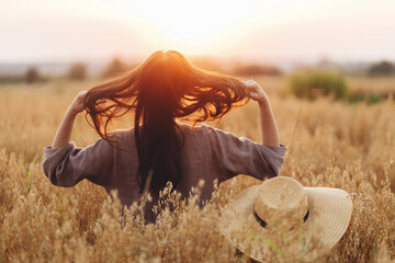 Stylish woman with straw hat standing in oat field in sunset light. Atmospheric tranquil moment....